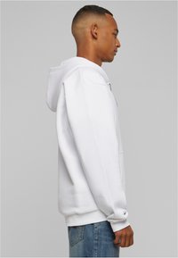 White hooded sweatshirt with a front pocket, ribbed cuffs, and hem. Model wears light blue jeans. Simple, casual design.