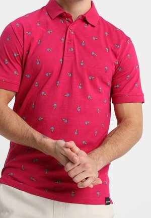 Homme portant un polo rouge avec un motif de petits cônes de glace bleus et marron clair, mains jointes devant.