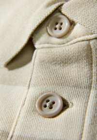 Beiges Strickgewebe mit einer gerippten Textur, mit zwei runden, vierlochigen Knöpfen in einem helleren Farbton, die auf einem Kragenabschnitt angebracht sind.