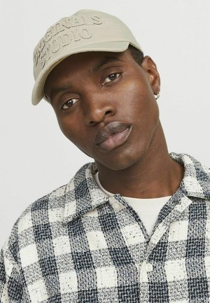 Beige cap with embossed "ORIGINALS STUDIO" text. Model wears a checkered blue and white textured shirt over a white top.