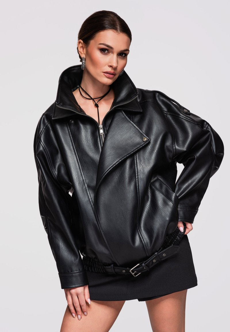 Vrouw draagt een oversized zwarte leren jas, korte zwarte rok, oorbellen en een zwarte halsband, poseert met hand op heup.