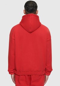 Sweatshirt à capuche rouge avec une coupe ample et des manches longues. Présente des poignets et un ourlet côtelés, ainsi qu'une texture de tissu lisse. Aucune logo ni motif visible.