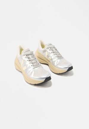 Hellbeige-weiße Laufschuhe mit dicken Sohlen, grauen Schnürsenkeln, perforierter Spitze und Logo an Seite und Zunge, auf weißem Hintergrund positioniert.