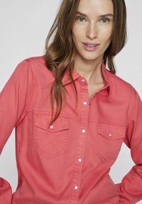 Camicia coral di cotone, maniche lunghe, due tasche sul petto con pattina, tessuto leggero, texture morbida e dettagli di cucitura visibili.