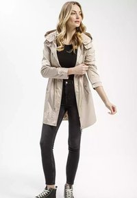 Jasnobeżowy wodoodporny płaszcz z kapturem na sznurku i zamkiem błyskawicznym, połączony z czarnymi dopasowanymi dżinsami i czarnymi wysokimi trampkami.