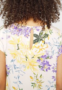 Bloemenjurk met korte mouwen, met gele en paarse bloemen op een lichte achtergrond, sleutelgatdetail aan de achterkant met een knoopsluiting.