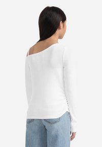 Bela dolga majica z dolgimi rokavi, širokim odprtim ovratnikom, rebrasto teksturo in gubanjem na hrbtu, v kombinaciji s svetlo modrimi kavbojkami.