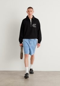 Felpa nera con zip e logo "CK", abbinata a pantaloni shorts in denim blu chiaro. Accentuata da calze bianche e mocassini neri, mentre porta una borsa marrone.