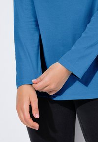 Niebieska koszula z długim rękawem o luźnym kroju, z miękką teksturą i bocznymi rozcięciami. Ręka poprawia mankiet przy czarnych spodniach.