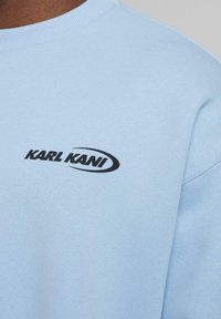 Gaiši zils kokvilnas sporta krekls ar melnu "KARL KANI" logo uz krūšu. Apģērba stils ir ar apaļu kakla izgriezumu un ribota apdare.