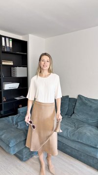 Lysebeige bluse med løse ærmer, tan midiskjørt og matchende hæler. Holder solbriller og en brun stropp. Teksturert blå sofa i bakgrunnen.