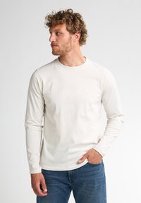 Camiseta de manga larga color crema hecha de algodón suave, con texto en relieve sutil en el pecho, combinada con jeans de mezclilla azules.