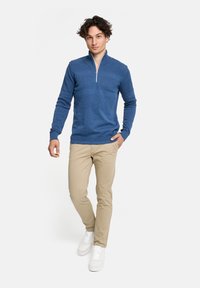 Blauer Zip-Hals-Pullover mit strukturiertem Muster, langen Ärmeln und geripptem Saum, kombiniert mit beigen Hosen und weißen Sneakers.