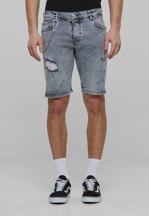 Shorts en denim gris délavé avec des détails usés, un accessoire en chaîne accroché et un ourlet roulé. Associés à des baskets noires et des chaussettes blanches.