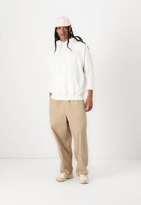 Sudadera blanca con capucha y bolsillo frontal, combinada con pantalones beige sueltos y zapatillas beige, llevada con una gorra de color claro.
