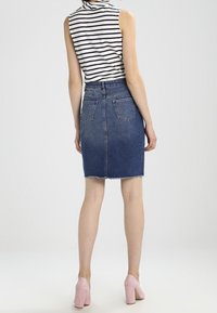 Denim kokerrok met rafelrand, blauwe kleur, en twee achterzakken, gecombineerd met een gestreept mouwloos topje en roze hoge hakken.