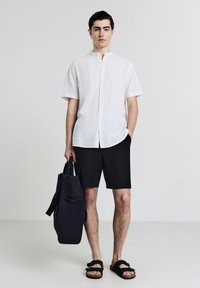 Mann in gestreiftem kurzärmeligen Hemd, schwarzen Shorts und schwarzen Sandalen, der eine große schwarze Tragetasche hält und vor einem einfarbigen hellen Hintergrund steht.