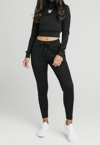 Černá krátká mikina s kapucí a logem, doplněná černými úzkými tepláky s elastickým pasem. Outfit doplňují bílé tenisky.