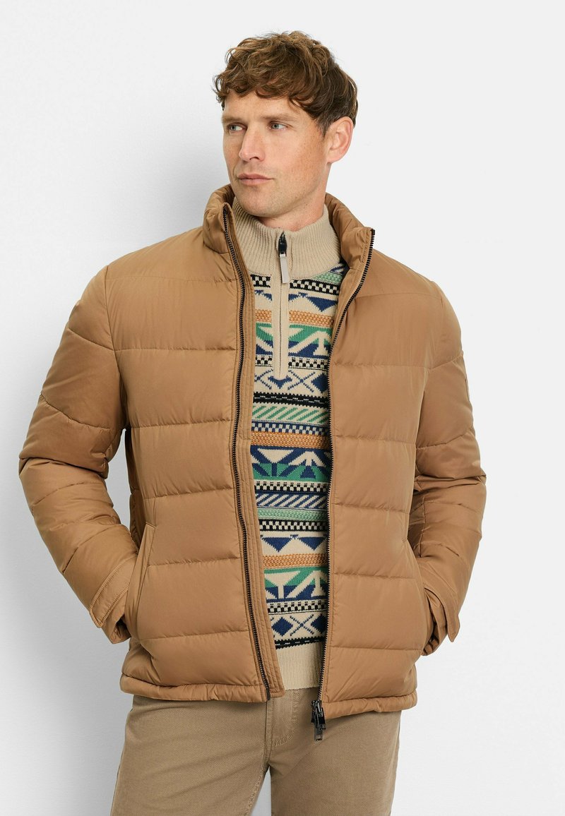 Helle braune Daunenjacke mit hohem Kragen und gestepptem Design, über einem gemusterten Pullover mit geometrischen Formen in Grün, Blau und Beige getragen.
