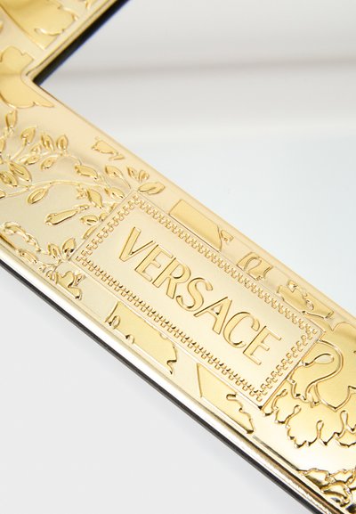 Złota ramka lustra Versace z misternie wytłaczanymi kwiatowymi wzorami oraz wypukłą tabliczką z logo i nazwą marki. Elegancka czarna krawędź.