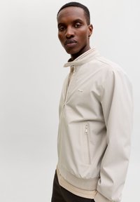 Ανοιχτό μπεζ μπουφάν με ψηλό γιακά, φερμουάρ μπροστά και ριμπ τελείωμα. Διαθέτει τσέπη με φερμουάρ και διακριτική λεπτομέρεια λογότυπου στο στήθος.