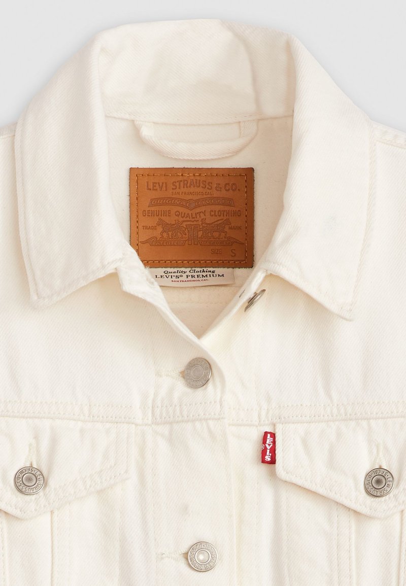 Chaqueta de mezclilla blanca con cuello puntiagudo, botones a presión plateados y una etiqueta de marca de cuero con detalles en relieve en el interior del cuello.