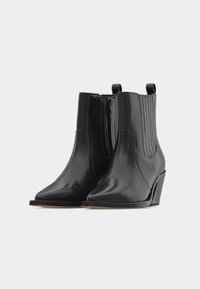 Ζευγάρι κομψών μαύρων δερμάτινων μποτάκια αστραγάλου με μυτερές μύτες, πλαϊνές φερμουάρ, ελαστικές επενδύσεις και τακούνια σφήνας σε απλό φωτεινό φόντο.