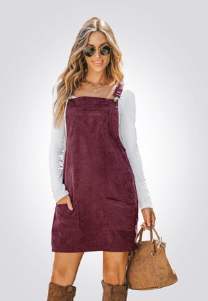 Corduroy Latzkleid in Dunkelviolett mit zwei Fronttaschen, verstellbaren Trägern und geradem Schnitt. Das Model hält eine braune Tasche und trägt ein weißes Oberteil.