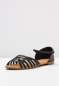 Black flat sandal featuring open-toe design with cut-out stripes, ankle strap, and brown sole. Made from smooth, matte material.