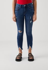 Τζιν παντελόνι σε σκούρο μπλε τζιν με φθαρμένα σκισίματα, φθαρμένες άκρες και ψηλή μέση. Συνδυασμένο με μαύρες σαγιονάρες με λουριά.