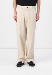 Beige, wijde broek met een textuurafwerking, voorzien van voorzakken en een knoopsluiting, gecombineerd met zwarte sneakers.