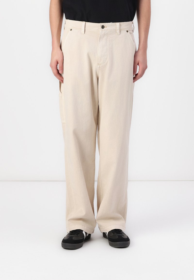 Beige, wijde broek met een textuurafwerking, voorzien van voorzakken en een knoopsluiting, gecombineerd met zwarte sneakers.
