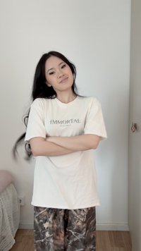 Junge Frau mit langen schwarzen Haaren verschränkt die Arme, trägt ein weißes "IMMORTAL"-T-Shirt und gemusterte Hosen, steht in einem minimalistischen Raum.
