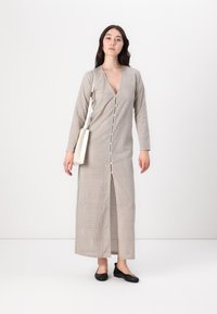 Dlhé béžové šaty s výstrihom do V, zapínaním na gombíky vpredu a dlhými rukávmi, ktoré majú jemný bodkovaný vzor. Styled s čiernymi balerínkami a taškou.