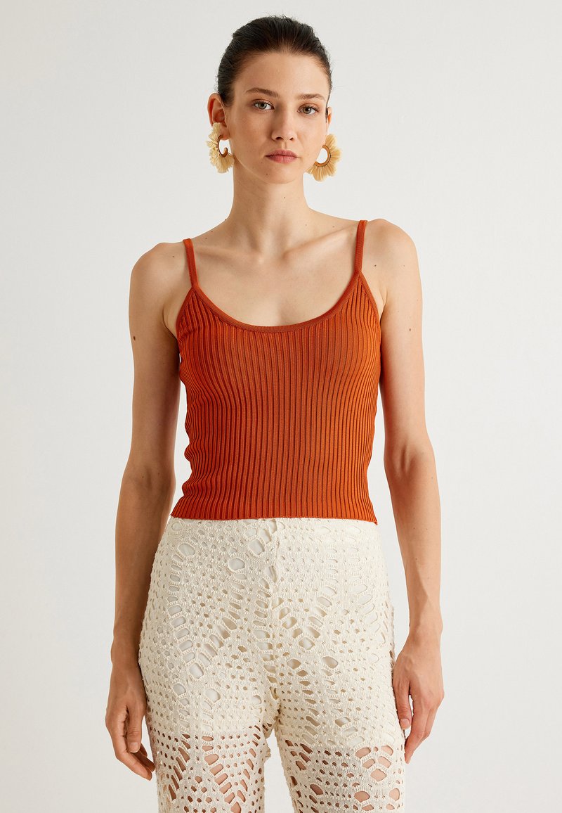 Canotta aderente a coste in arancione bruciatore con spalline sottili. Abbinata a pantaloni in pizzo bianco intricati con motivi a ritaglio.