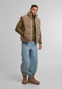 Barna fleece mellény olívazöld hosszúujjú póló felett, világoskék, bő szabású farmernadrággal és bézs fűzős csizmával párosítva. Sima textúra, laza fazon.