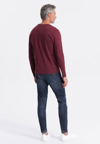 Camisa de manga larga color burdeos con cuello redondo, combinada con jeans ajustados de color azul y zapatillas blancas. Textura suave, diseño casual y con un mínimo de detalles.