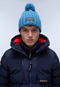 Beanie azzurro lavorato a maglia con pon-pon in cima, caratterizzato da un motivo testurizzato. Indossato con un giubbotto imbottito blu navy, impreziosito da dettagli rossi.