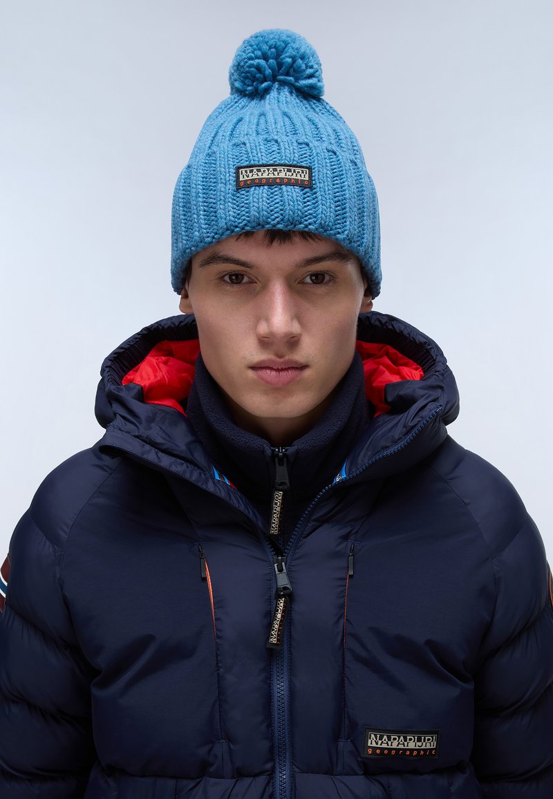 Beanie azzurro lavorato a maglia con pon-pon in cima, caratterizzato da un motivo testurizzato. Indossato con un giubbotto imbottito blu navy, impreziosito da dettagli rossi.