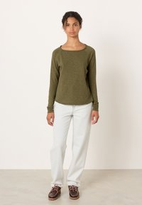 Noor naine seisab, kandes oliivrohelist pika varrukaga pusa, helerohelisi teksapükse ja pruune paeltega kingi tavalise hele tausta ees.