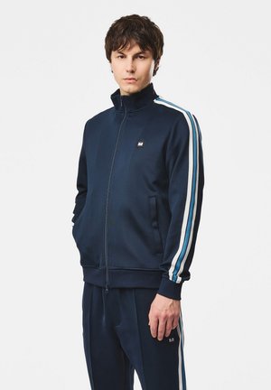 Jeune homme portant un survêtement bleu marine avec des rayures blanches et bleu clair sur les manches et le pantalon, debout devant un fond blanc uni.