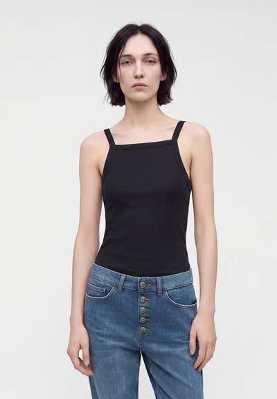 Jeune femme aux cheveux courts et foncés, portant un haut noir sans manches et un jean bleu avec plusieurs boutons, debout devant un fond uni.