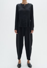 Svart, satin långärmad skjorta med krage som knäpps och löst sittande byxor, med smalnande anklar och platta svarta skor.