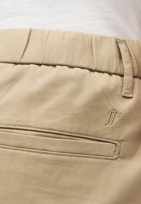 Μπεζ παντελόνια με ελαστική ζώνη μέσης, πίσω τσέπη με περιμετρικό τελείωμα, θηλιά για ζώνη και μικρό κεντητό λογότυπο κοντά στην τσέπη σε ανοιχτό ύφασμα.