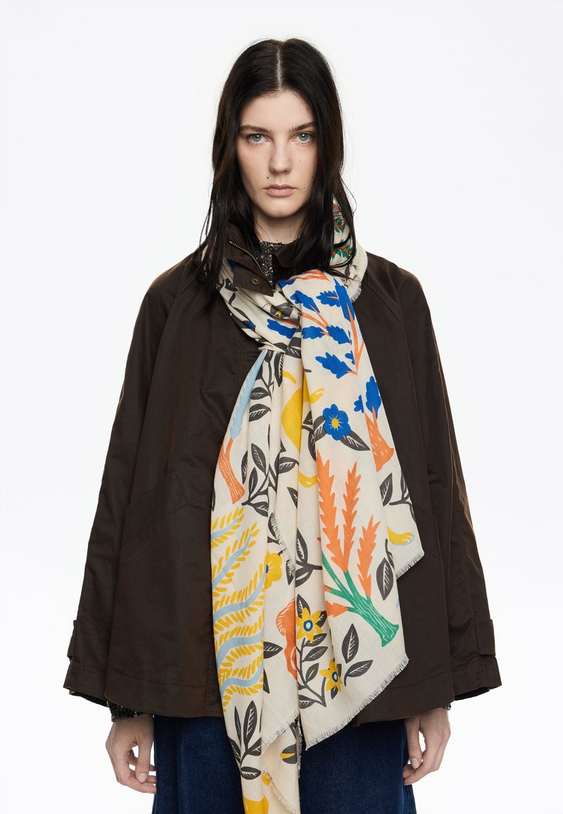 Gran bufanda floral que presenta patrones en azul, naranja y amarillo sobre un fondo crema, drapeada sobre una chaqueta exterior marrón. Textura suave.