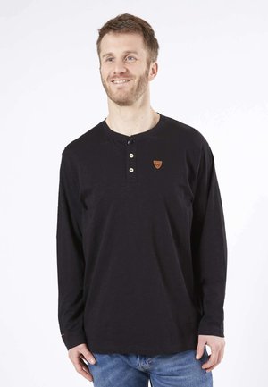 Chemise noire à manches longues avec un col rond, trois boutons et un petit patch logo marron sur la poitrine. Texture douce, coupe décontractée.