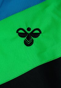 Barevná látková nášivka se zelenými, černými a modrými sekcemi; černé vyšívané logo připomínající stylizovaný hmyz nebo abstraktní design.