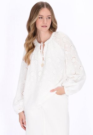 Fiatal nő hosszú, hullámos hajjal, fehér, hosszú ujjú, fűzős nyakú hímzett blúzban és fehér nadrágban, oldalra nézve.