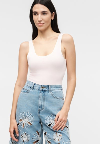 Γυναίκα με μακριά σκούρα μαλλιά φοράει άσπρη κοντή μπλούζα και ανοιχτό μπλε τζιν με ψηλή μέση και μεγάλα λουλουδένια κοψίματα στα πόδια.