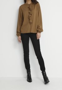 Camicia marrone a maniche lunghe con dettagli arricciati sul davanti, polsini arricciati, abbinata a pantaloni neri slim-fit e stivaletti.